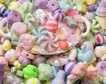 Puede incluir: Una colección de cuentas y abalorios de plástico de color pastel, incluyendo cuentas con forma de caramelo, corazones, estrellas y otras formas. Las cuentas están dispuestas en un cuenco y sobre una mesa.