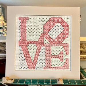 May include: A square cross-stitch artwork framed in a white mat, featuring the word LOVE in large red block letters with intricate geometric patterns. The background consists of a repeating pattern of small green and blue V-shaped stitches.