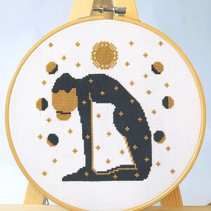 May include: A black and gold cross-stitch embroidery hoop with a design of a person in a yoga pose with a full moon and crescent moons in the background.