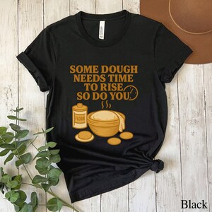 May include: Black t-shirt with a graphic design featuring a flour container, a bowl of dough, and the text "SOME DOUGH NEEDS TIME TO RISE SO DO YOU." The design is in shades of brown and yellow.