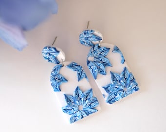 May include: A pair of blue and white floral dangle earrings. The earrings are made of polymer clay and have a white background with blue floral designs.