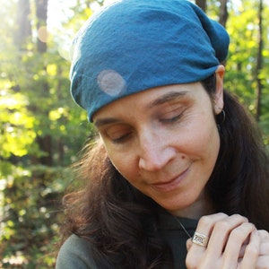 May include: A woman with long dark hair wearing a teal blue bandana and a green hooded sweatshirt. She is looking down at a silver ring on her finger.