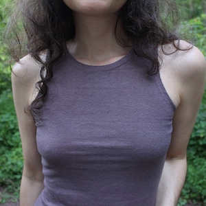 May include: A woman wearing a sleeveless, dark grey tank top. The top is made of a soft, textured fabric. The woman is standing in a woodland setting.