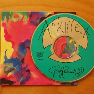 Può includere: Un disco compatto turchese con un centro dorato e un design verde e oro. Il disco ha il testo "Arkitex" e "Pure Research" scritto su di esso. Il disco è sopra una stampa d'arte astratta colorata.