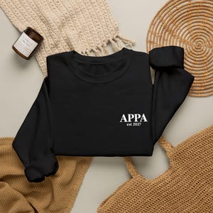 May include: A black long-sleeve sweatshirt featuring the white text APPA est 2027 printed on the front. The garment has a crew neckline, long sleeves with ribbed cuffs, and a solid black fabric construction with a clean, simple design.