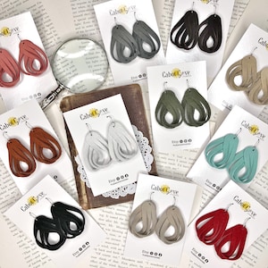 May include: Assortment of handmade leather earrings in various colors, including red, green, blue, brown, gray, and beige. The earrings are displayed on white cards with the text "Cabot Cove Creations" and "Etsy" printed on them.
