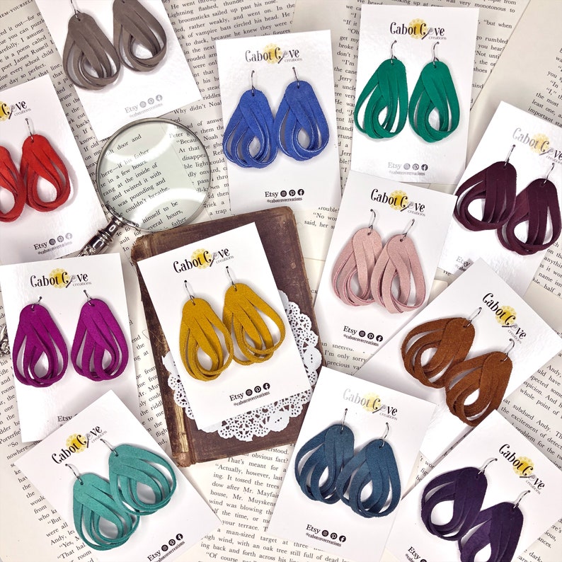May include: A collection of 10 pairs of dangle earrings made from suede leather. The earrings are displayed on white cards with the brand name "Cabot Cove Creations" printed on them. The earrings are in various colors, including purple, teal, brown, pink, blue, green, and gold.