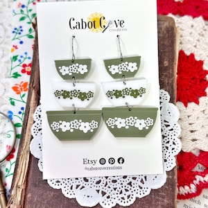May include: A pair of green and white ceramic bowl earrings with floral designs. The earrings are hanging from silver hooks. The earrings are displayed on a white card with the text "Cabot Cove creations" and "Etsy @cabotcovecreations".