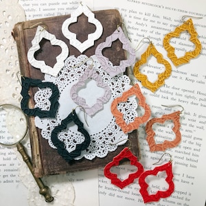 May include: Assortment of eight pairs of handmade leather earrings in various colors, including white, black, red, orange, and gold. The earrings are all in a quatrefoil shape with a textured surface. The earrings are displayed on a white doily and a vintage book.