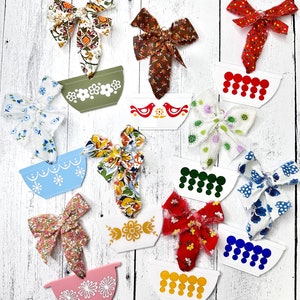 May include: A collection of 10 white ceramic bowls with various colorful designs. Each bowl has a different pattern, including birds, flowers, and geometric shapes. The bowls are decorated with red, blue, green, yellow, and brown fabric bows.