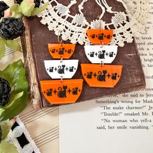 May include: A pair of orange and white layered cat-themed earrings. Each earring has three layers, with the top and bottom layers being orange and the middle layer being white. Each layer features a black silhouette of a cat with a tail.