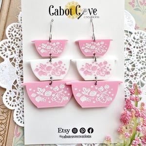 May include: Pink and white bowl-shaped earrings with floral designs. The earrings are made of acrylic and feature a layered design. The text "Cabot Cove creations" is at the top. The Etsy shop's social media icons are at the bottom.