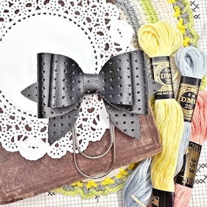 May include: A black leather bow with a dotted pattern, attached to a silver metal binder clip. The bow is sitting on a white doily and a brown surface. There are three spools of embroidery floss in the background, one yellow, one blue, and one pink.