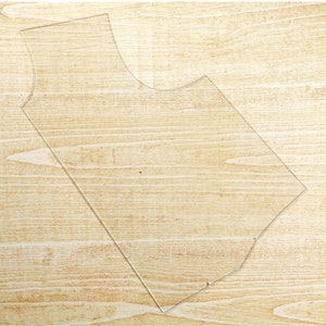 May include: A clear plastic template for sewing a garment, with a rounded neckline and curved bottom hem. The template is lying on a light brown wooden surface.