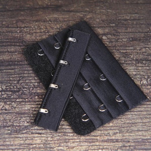 May include: Two black bra extenders with metal hooks and loops. The extenders are made of a soft, stretchy fabric and have a hook and loop closure.