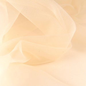 May include: A close-up of a soft, pale peach-colored tulle fabric. The fabric is draped and folded, creating a delicate and airy texture.