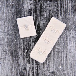 May include: Two beige fabric bra strap extenders with metal hooks. The extender on the left has one hook, and the extender on the right has three hooks.