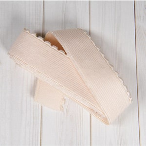 May include: A close-up of a beige elastic trim with a scalloped edge. The trim is folded in half and is lying on a white wooden surface.