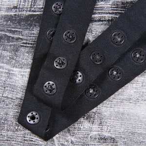 May include: Black elastic straps with multiple rows of black plastic buttons. The straps are folded in half and the buttons are visible on both sides.
