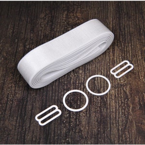 May include: A roll of white elastic ribbon and several white bra-making hardware pieces, including rings and sliders, are arranged on a dark wood surface. The components are for lingerie or garment construction.