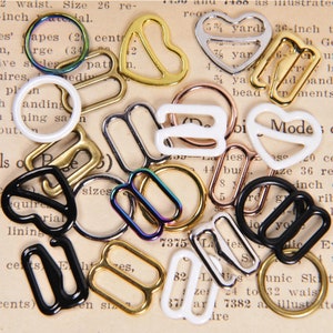 May include: Assortment of metal and plastic bra strap sliders in various shapes and colors, including gold, silver, black, white, and iridescent. Some sliders are heart-shaped, while others are rectangular or oval.