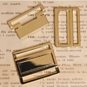 May include: Three gold-colored metal buckles in various shapes. The buckles have a shiny finish and are likely for use in clothing or accessories. The background is a vintage paper with text.