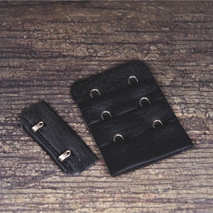May include: Two black bra extenders. One extender has two hook-and-eye closures, and the other has three hook-and-eye closures. The extenders are made of a woven fabric and are designed to add extra length to a bra band.