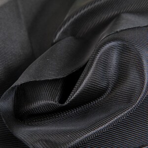 May include: Close-up of black fabric with a textured weave. The fabric is folded and draped, creating shadows and highlights that emphasize its texture. The material appears to be a durable, possibly water-resistant textile.