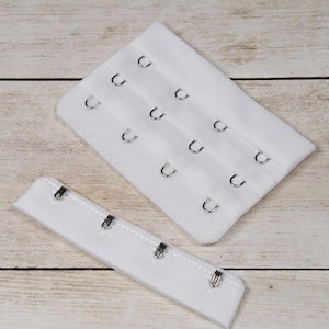 May include: Two white bra strap extenders with metal hooks. The larger extender has three rows of hooks, and the smaller extender has four hooks.