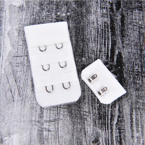 May include: White bra extender with three rows of hooks and eyes. The extender has a white fabric strap and metal hooks and eyes. The extender is shown on a dark wood background.