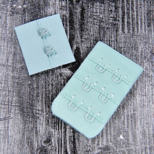 May include: Two light blue bra strap extenders with metal hooks. The extender on the left has three hooks and the extender on the right has three rows of hooks.