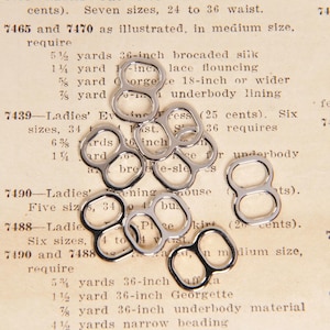 May include: A close-up of silver metal garment loops or rings. The loops are arranged in a pile on a vintage page of a sewing pattern book. The text on the page includes the numbers 7465, 7470, 7439, 7490, 7488, and the words "Ladies' Evening Dress", "Five sizes", "Six sizes", "require", "yards", "inch", "underbody", "material", "narrow", "beading".