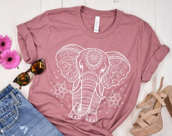 May include: A dusty rose t-shirt with a white mandala elephant design. The shirt is laid out on a white wooden surface with a pair of blue denim shorts, sunglasses, and a pair of beige heeled sandals.