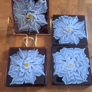 May include: Set of four square coasters with a dark brown background and a white flower design. The flowers have a light blue color and a yellow center.