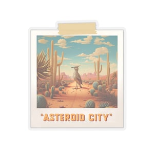 May include: A vintage-style photo of a desert landscape with a bird standing in the middle of a dirt road. The photo is framed with a white border and has a yellow tape across the top. The text "ASTEROID CITY" is printed below the photo.