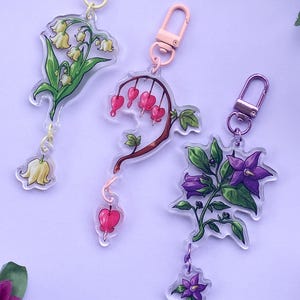 Può includere: Tre portachiavi in acrilico con disegni floreali. Uno raffigura mughetto con fiori gialli, un altro cuori sanguinanti e il terzo fiori viola e foglie verdi. Ogni portachiavi ha una chiusura colorata.