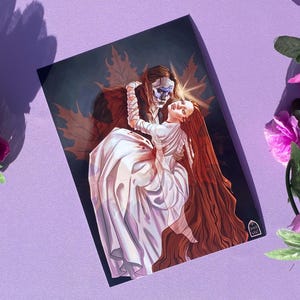 Puede incluir: Una impresión artística rectangular que presenta una escena gótica romántica. Una figura enmascarada con ropa oscura abraza a una mujer con un vestido blanco. La mujer tiene el pelo largo y rojo. El fondo es oscuro con hojas de otoño.