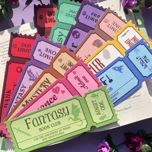Könnte beinhalten: Eine Sammlung farbenfroher Lesezeichen in Ticketform mit verschiedenen Designs und dem Schriftzug "Admit One". Die Lesezeichen liegen auf einem offenen Buch, auf einem ist "Fantasy Book Club" zu lesen. Die Farben sind Rot, Lila, Rosa und Grün.