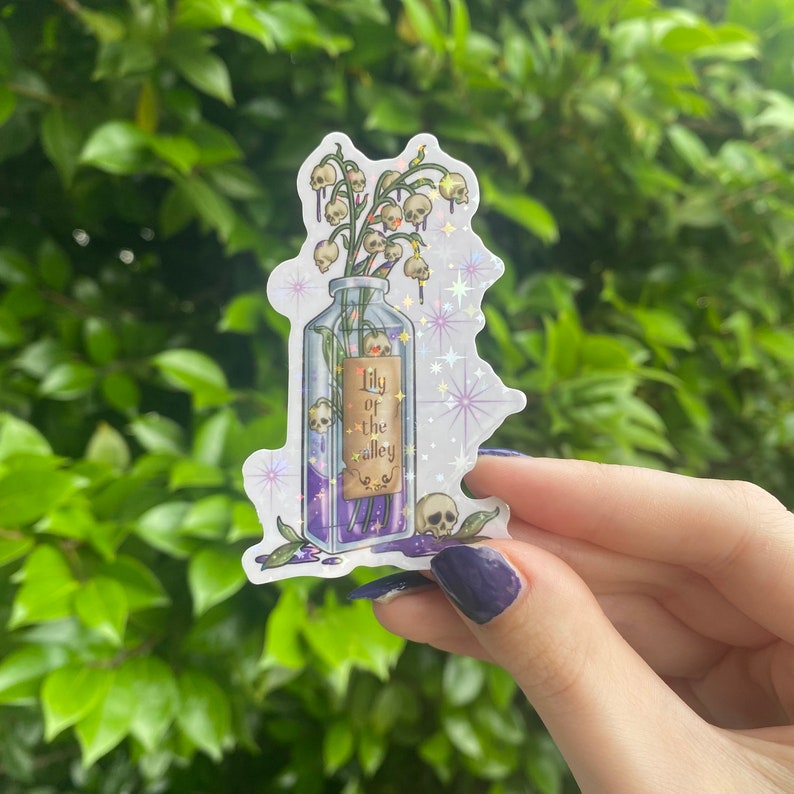 Puede incluir: Una botella de vidrio transparente llena de un l&iacute;quido morado y un ramo de flores blancas de lirio de los valles con calaveras como flores. La botella tiene una etiqueta que dice "Lily of the Valley". La pegatina tiene un fondo brillante.