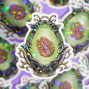 Pode incluir: Um adesivo holográfico de um abacate verde com um cérebro dentro. O abacate é rodeado por uma moldura dourada e preta com estrelas.