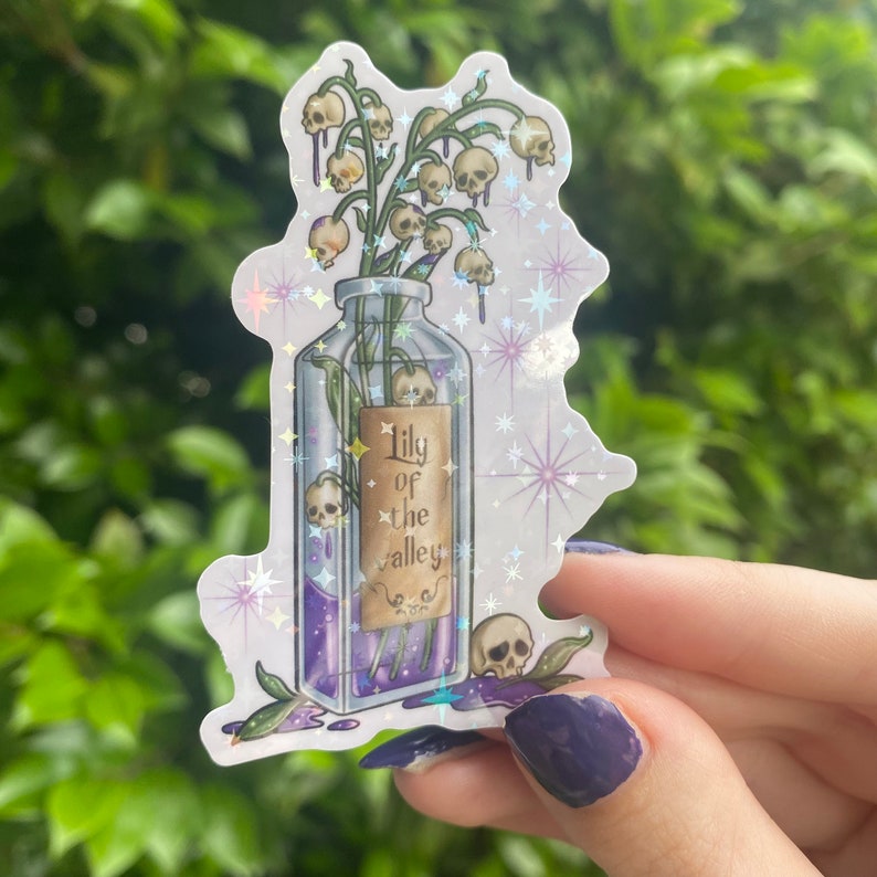 Puede incluir: Una botella de vidrio transparente con un l&iacute;quido morado y una etiqueta que dice "Lily of the Valley". La botella est&aacute; llena de un ramo de flores blancas con calaveras al final de cada tallo. La botella est&aacute; rodeada de un fondo brillante con estrellas.