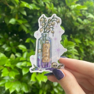 Puede incluir: Una botella de vidrio transparente llena de un l&iacute;quido morado y una etiqueta que dice "Lily of the Valley". La botella est&aacute; llena de flores blancas y calaveras. El fondo es blanco brillante.