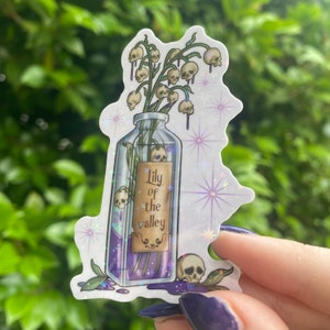 Puede incluir: Una botella de vidrio transparente llena de un l&iacute;quido morado y un ramo de lirios del valle con calaveras como flores. La botella tiene una etiqueta que dice "Lily of the valley".