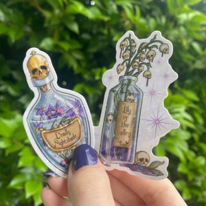 Puede incluir: Dos botellas de vidrio con etiquetas y flores moradas en su interior. La botella de la izquierda tiene un tap&oacute;n de cr&aacute;neo y una etiqueta que dice "Deadly Nightshade". La botella de la derecha tiene una etiqueta que dice "Lily of the Valley".