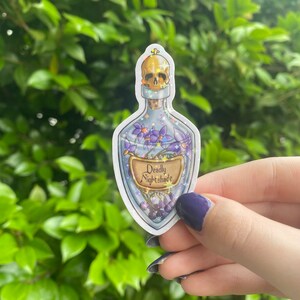 Puede incluir: Una pegatina de una botella de vidrio con un tap&oacute;n de calavera, llena de flores moradas y una etiqueta que dice "Deadly Nightshade".