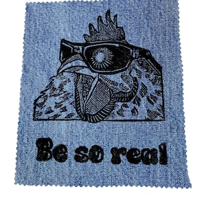 May include: A black and white illustration of a rooster wearing sunglasses and a crown on a blue denim background. The text "Be so real" is printed below the rooster.