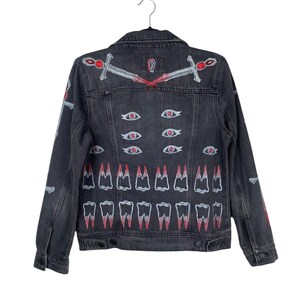 May include: A black denim jacket with hand-painted artwork. The back features white and red designs of swords, eyes, and teeth. The jacket has a classic collar, button closures, and long sleeves.
