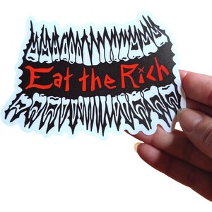May include: A sticker with a black and white design resembling teeth, with the words "Eat the Rich" in red. The sticker has a white border and is held by a hand.