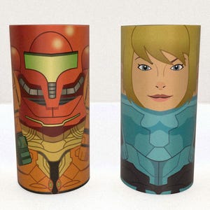 May include: Two cylindrical tumblers with cartoon illustrations. One features a red and orange armored figure with a green visor. The other depicts a blonde woman in a blue and black armored suit. Both tumblers are on a white surface.