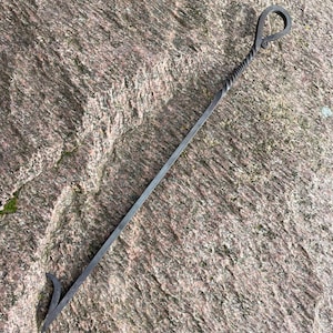 Könnte beinhalten: Ein langer, schwarzer Kaminbesen aus Metall mit einem gedrehten Griff und einem Haken am Ende. Der Besen liegt auf einer rauen, grauen Steinoberfläche.